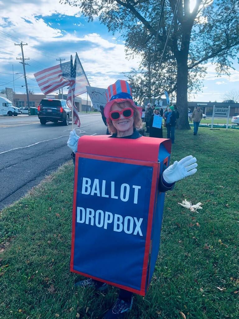 A woman in a homemade blue and red ballot drop box costume and Uncle Sam hat holds a small American flag